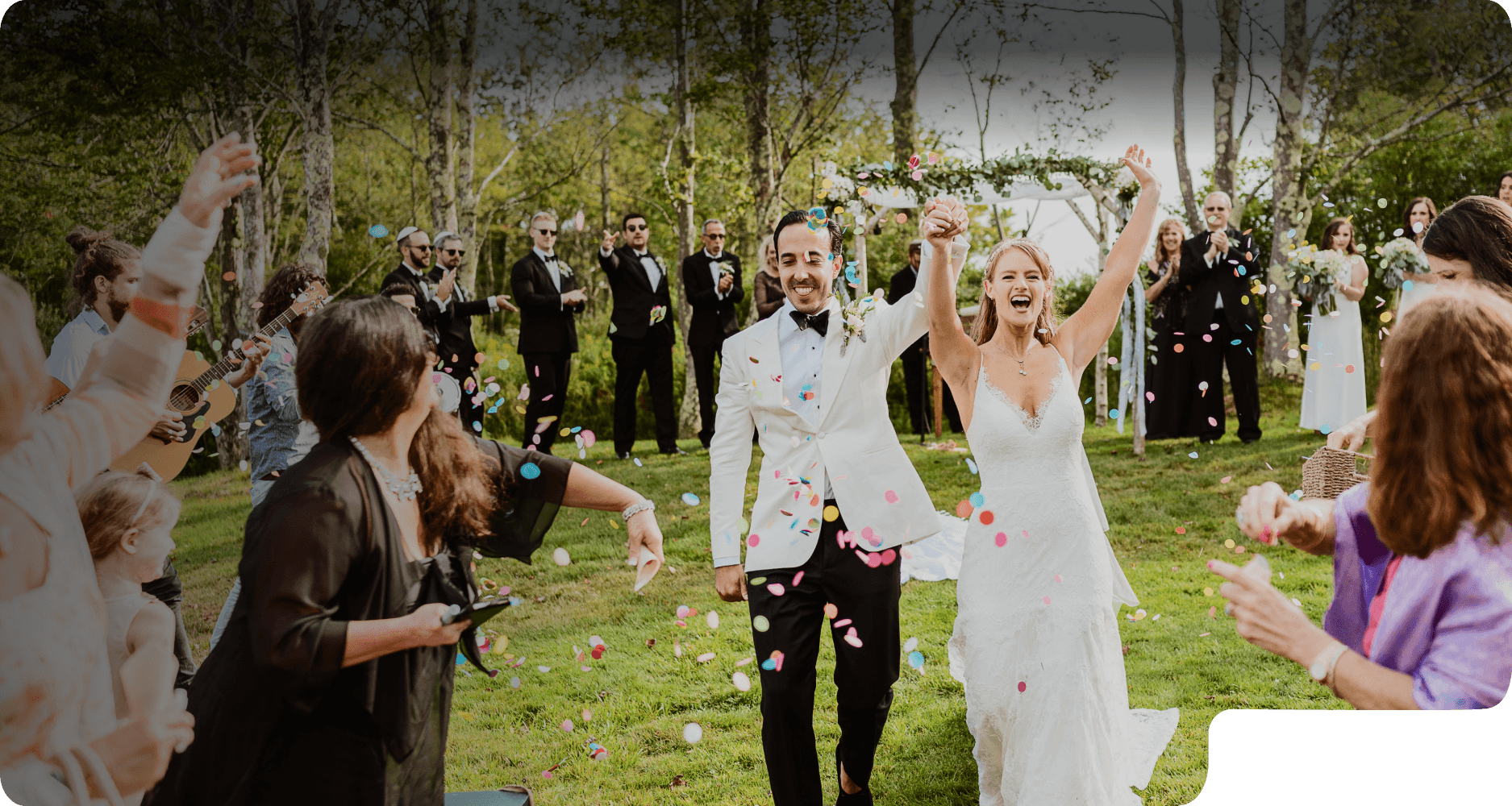 A bride and groom walking through a shower of confetti as their guests cheer at an outdoor Jewish wedding