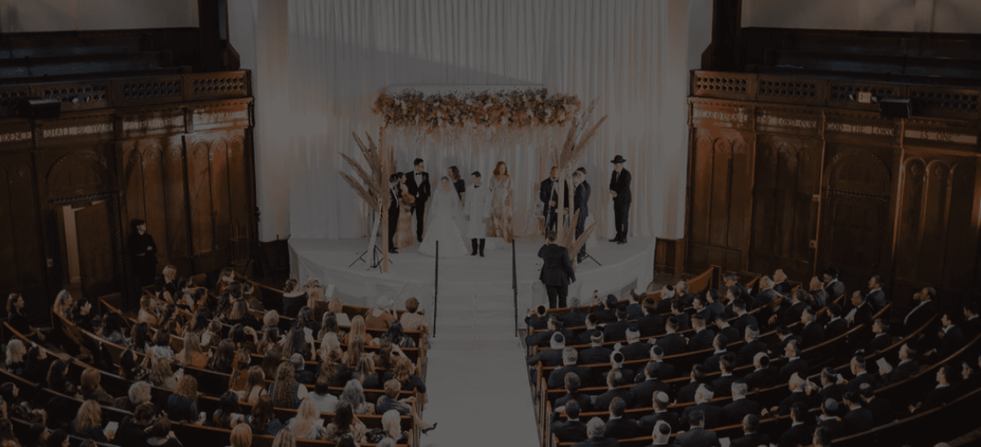 A wedding ceremony under a flower-topped chuppah in a grand hall, seen from above a full congregation