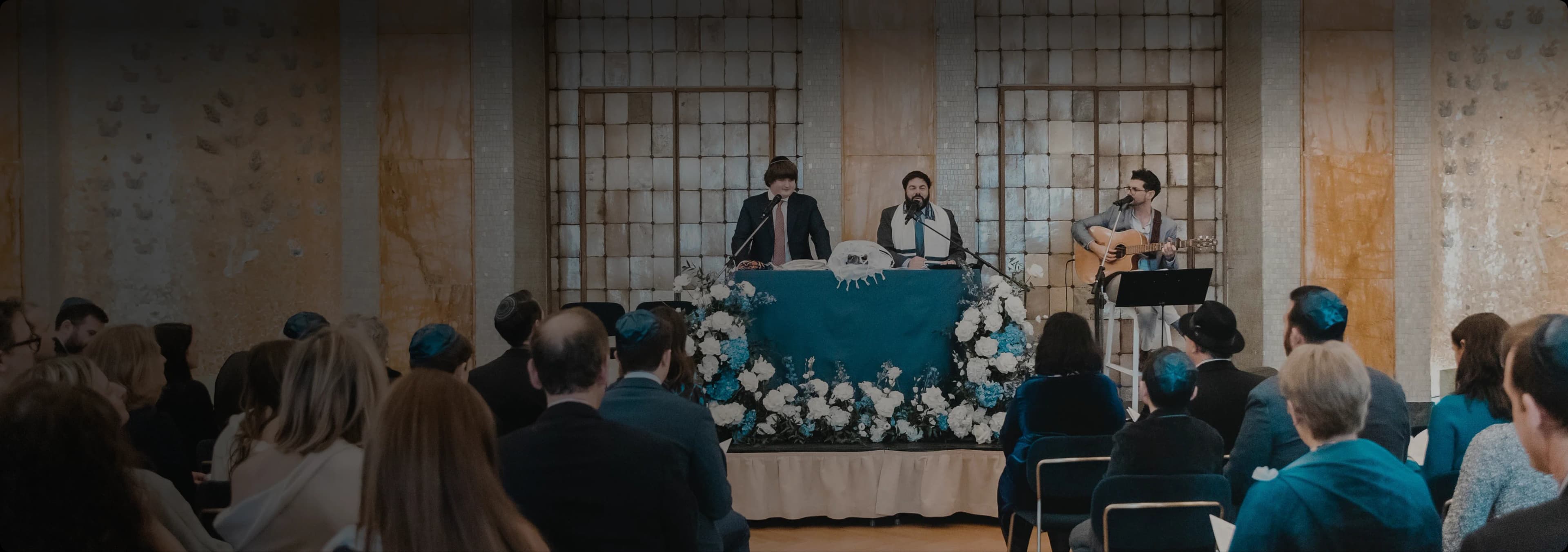 Three musicians performing beside a bimah dressed with blue and white flowers, the congregation seated in front