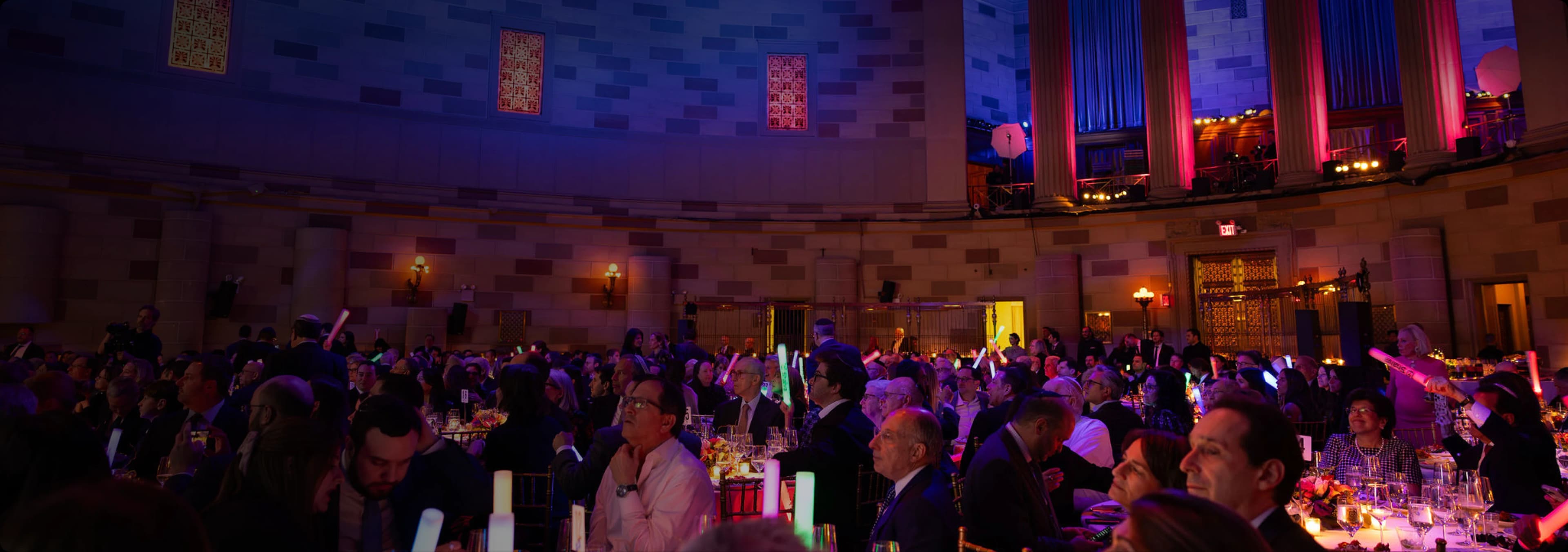 Guests at dinner tables filling a grand hall lit in blue and pink for a gala evening