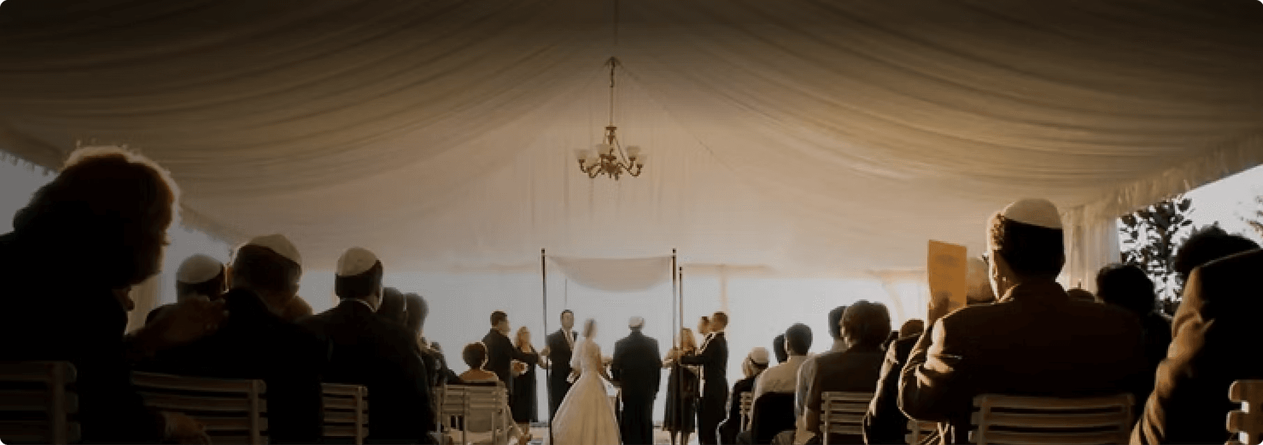 Guests seated under a white marquee watching a wedding ceremony at the chuppah