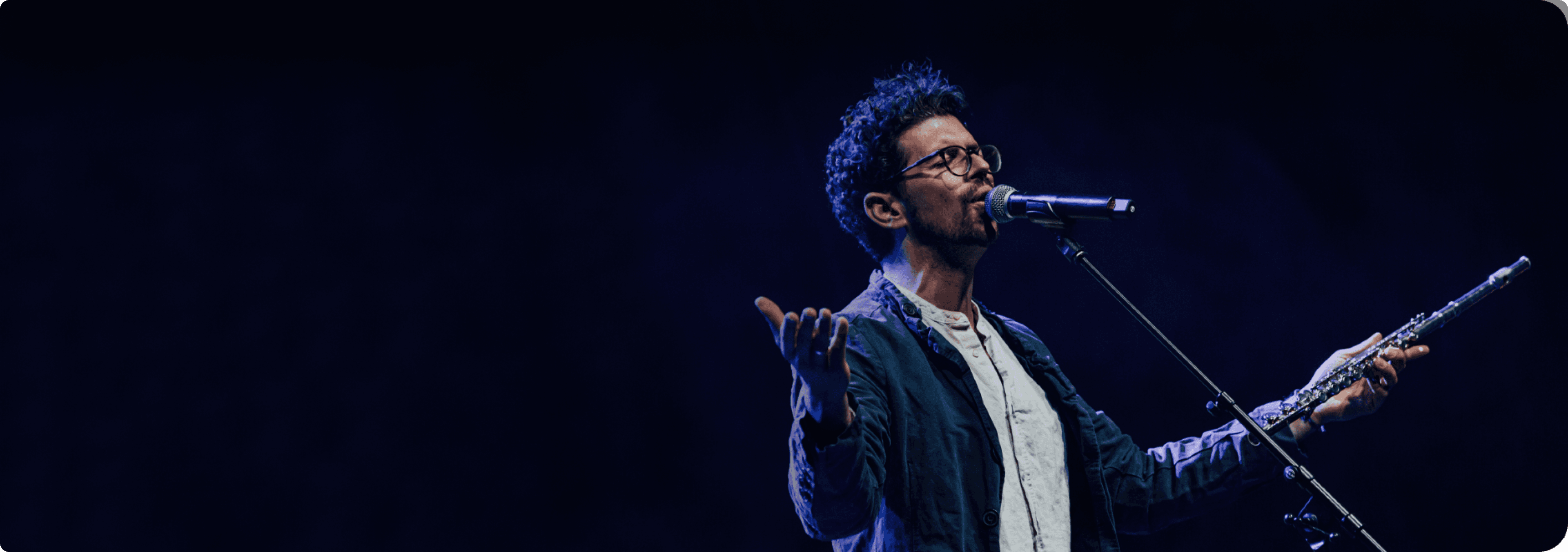 Yoni Avital singing into a microphone on a darkened stage, a flautist playing beside him